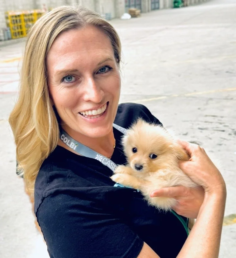 Blonde woman named alyssa christian wearing a black polo shirt and a light blue lanyard, smiling while holding a small fluffy tan puppy outdoors