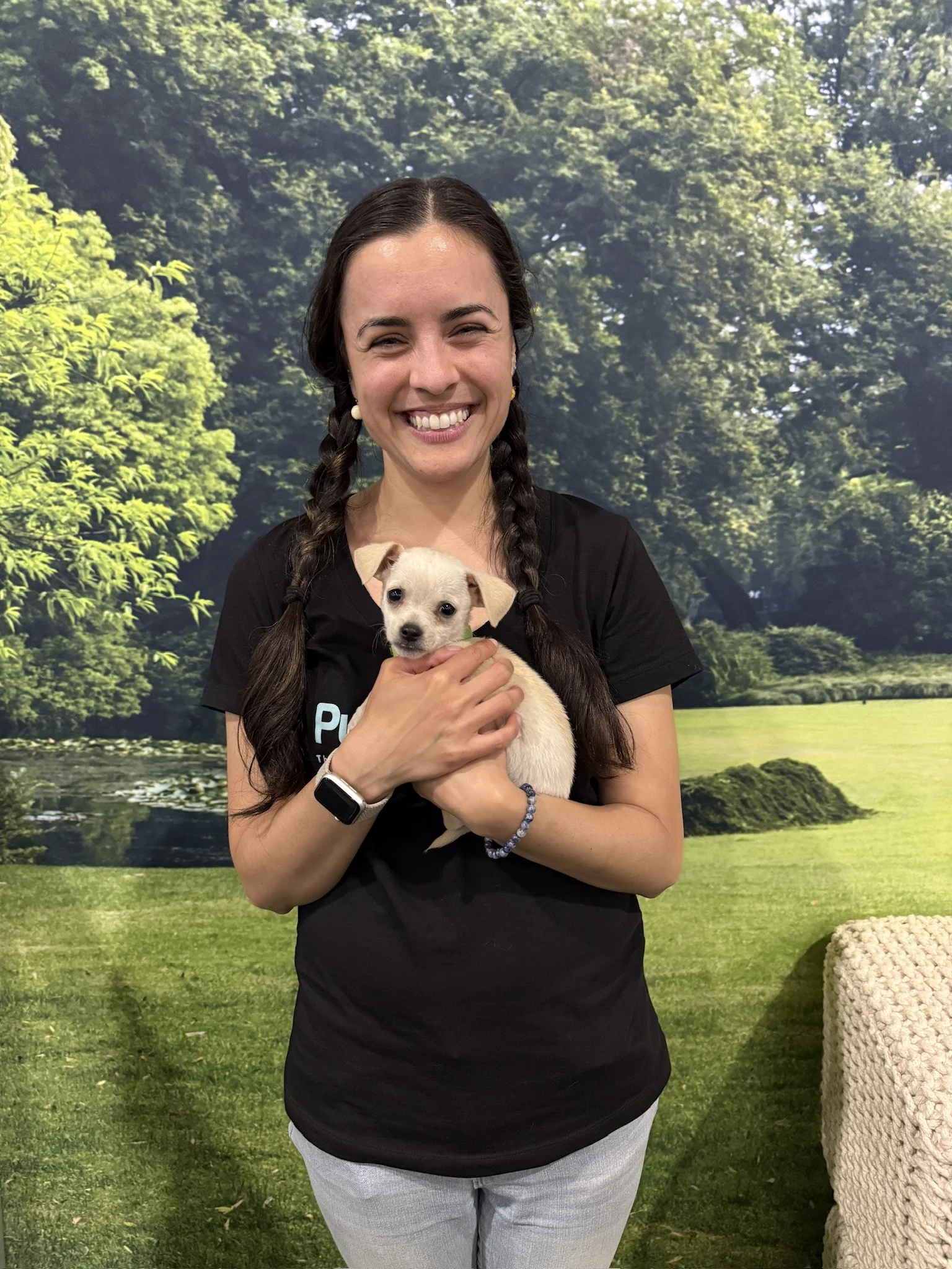 A smiling woman with braided dark hair wearing a black shirt and holding a small cream-colored chihuahua puppy outdoors.