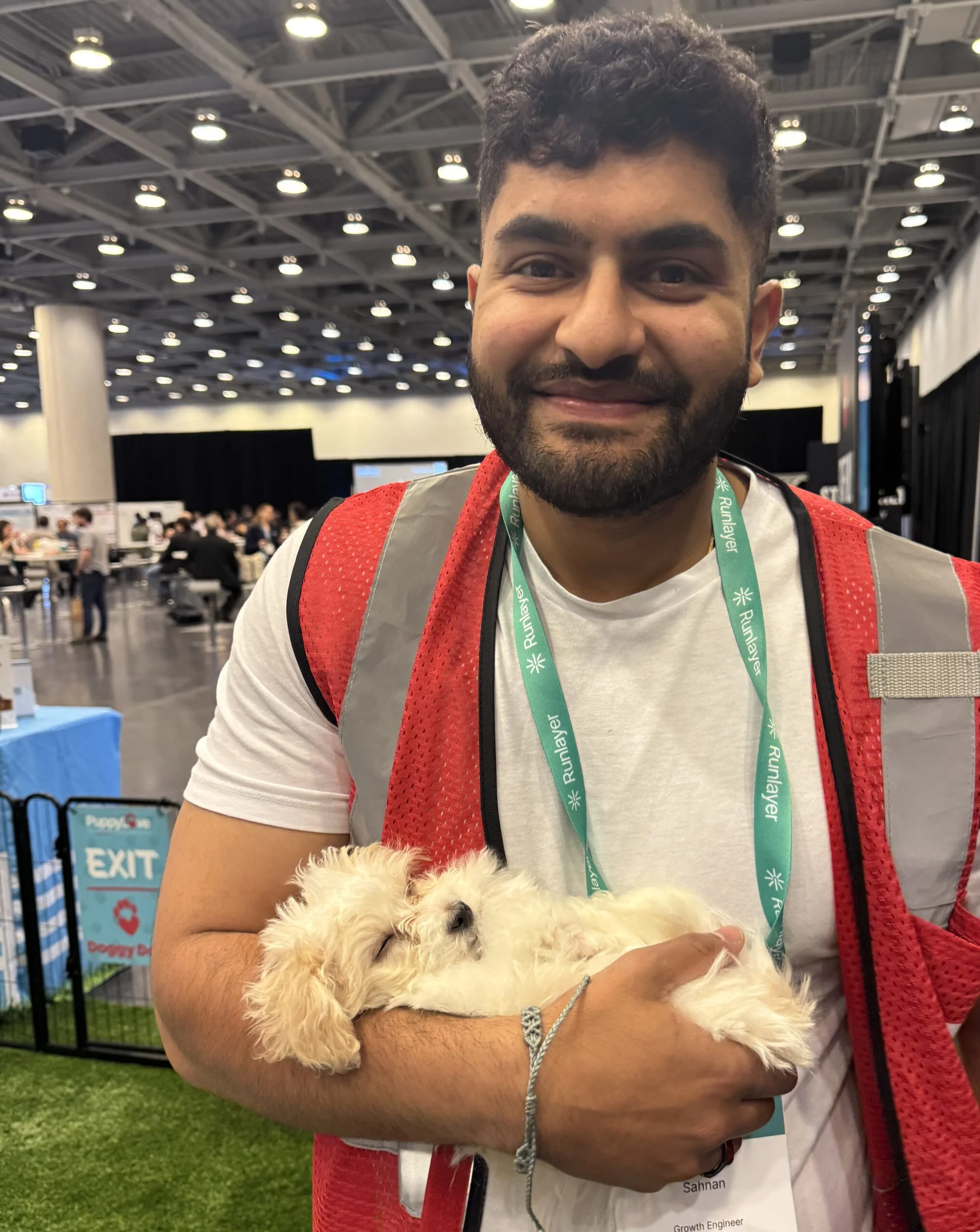 A smiling man with short dark hair and a beard wearing a white t-shirt and red safety vest, cradling a small fluffy white puppy in his arms indoors.