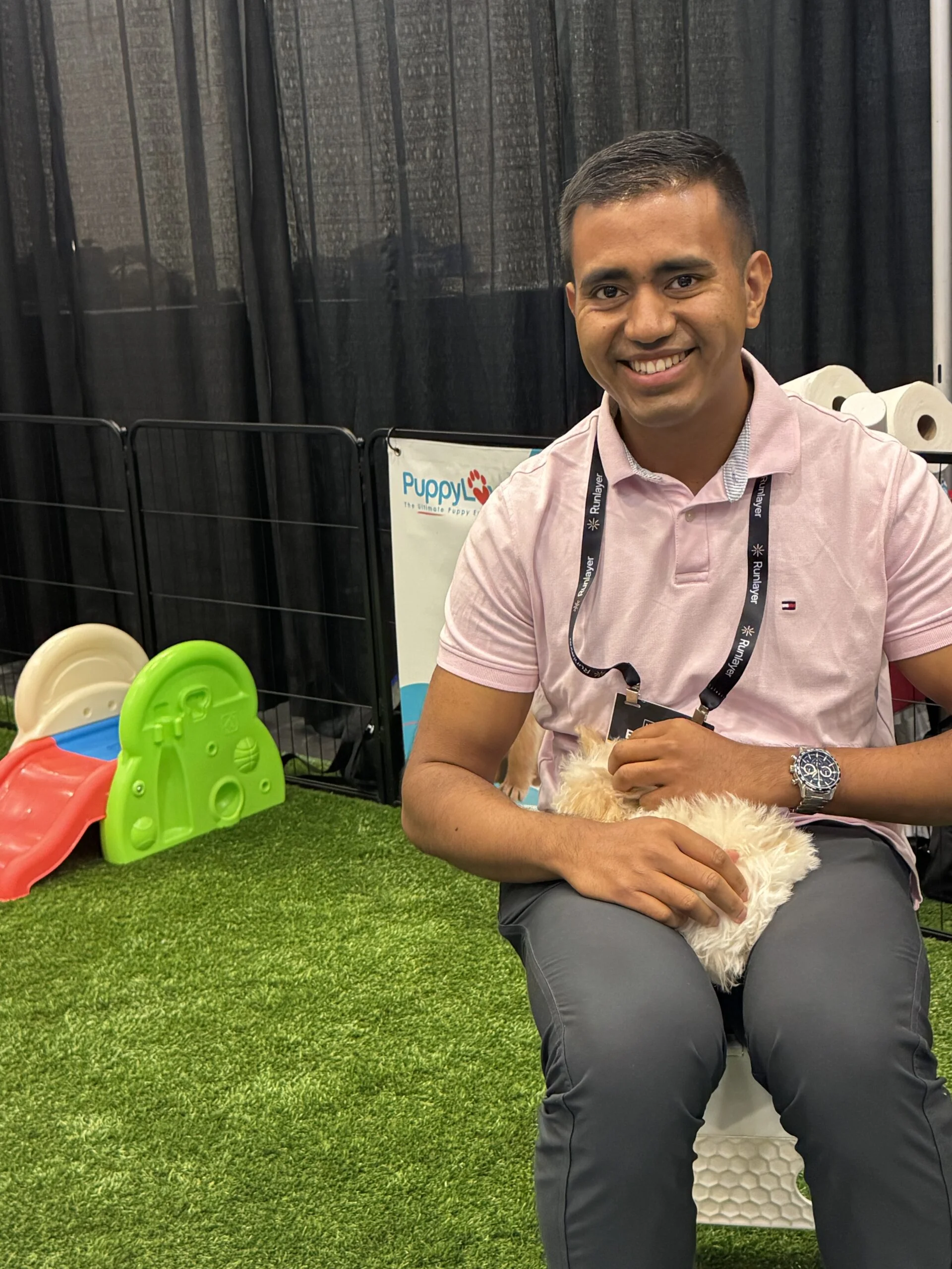 A smiling man with short dark hair wearing a pink polo shirt sitting on artificial turf while cradling a small fluffy white puppy in his lap indoors.