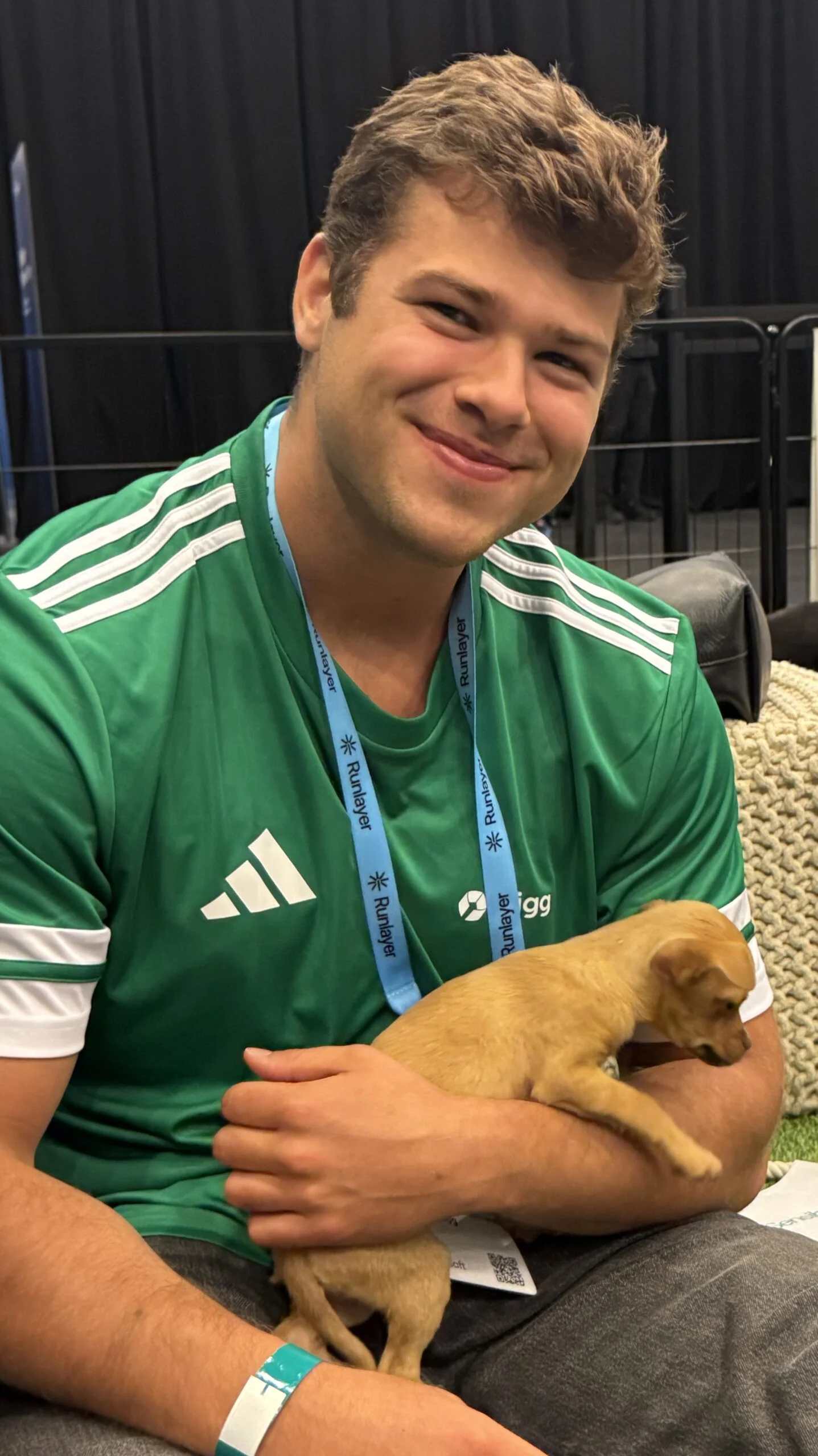 A smiling young man with short brown hair wearing a green and white striped athletic jersey, holding a small tan puppy in his lap indoors.