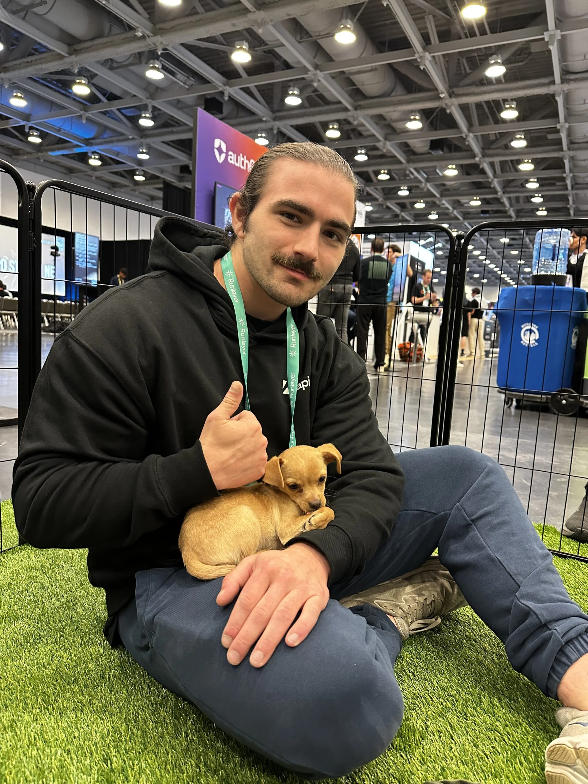 A smiling man with a mustache and hair bun wearing a black hoodie with a green lanyard, sitting on artificial turf while holding a small tan puppy in his lap and giving a thumbs-up gesture.