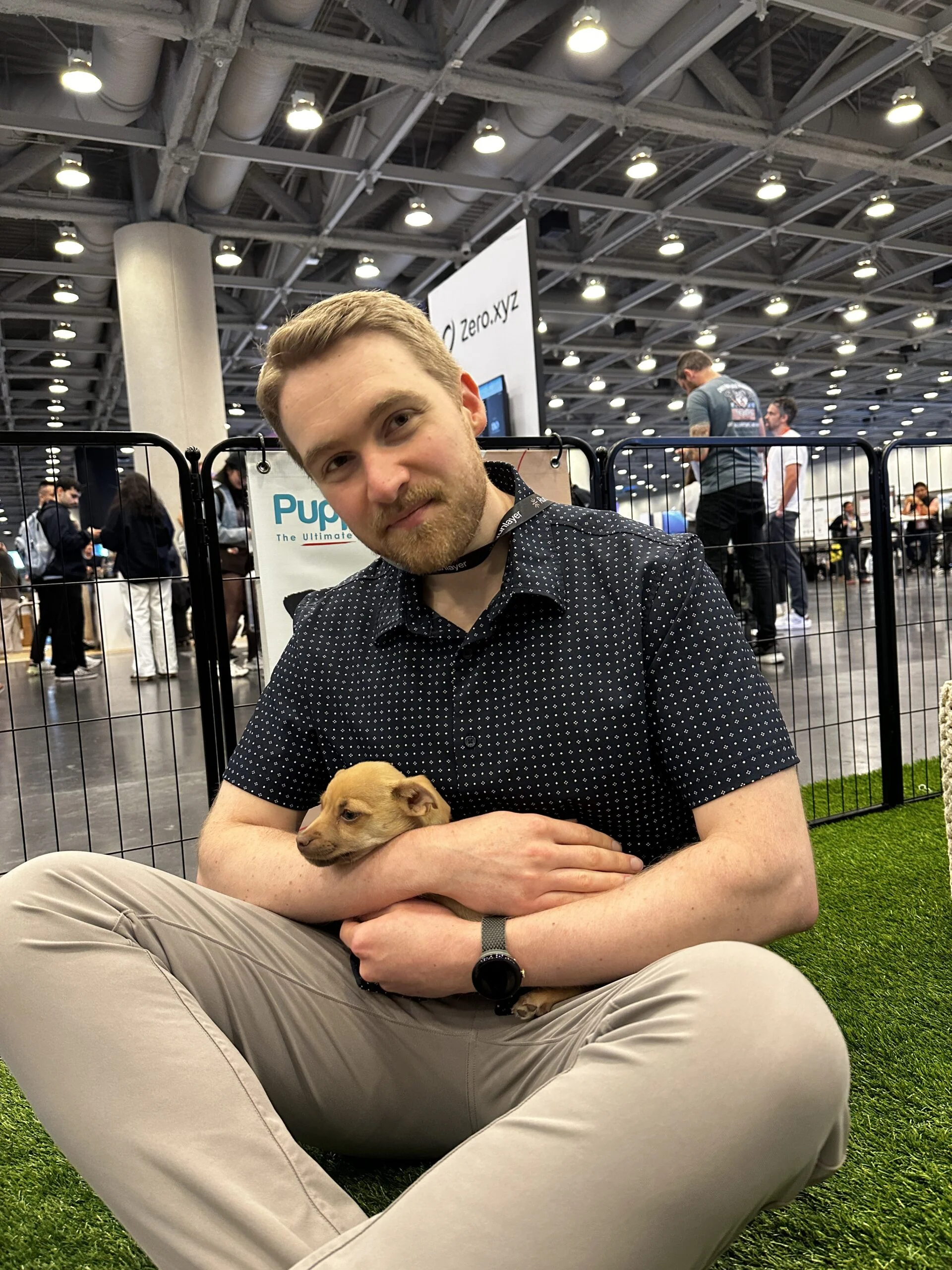 A smiling man with a short beard wearing a blue button-down shirt sitting on artificial turf while holding a small light-brown puppy in his arms.