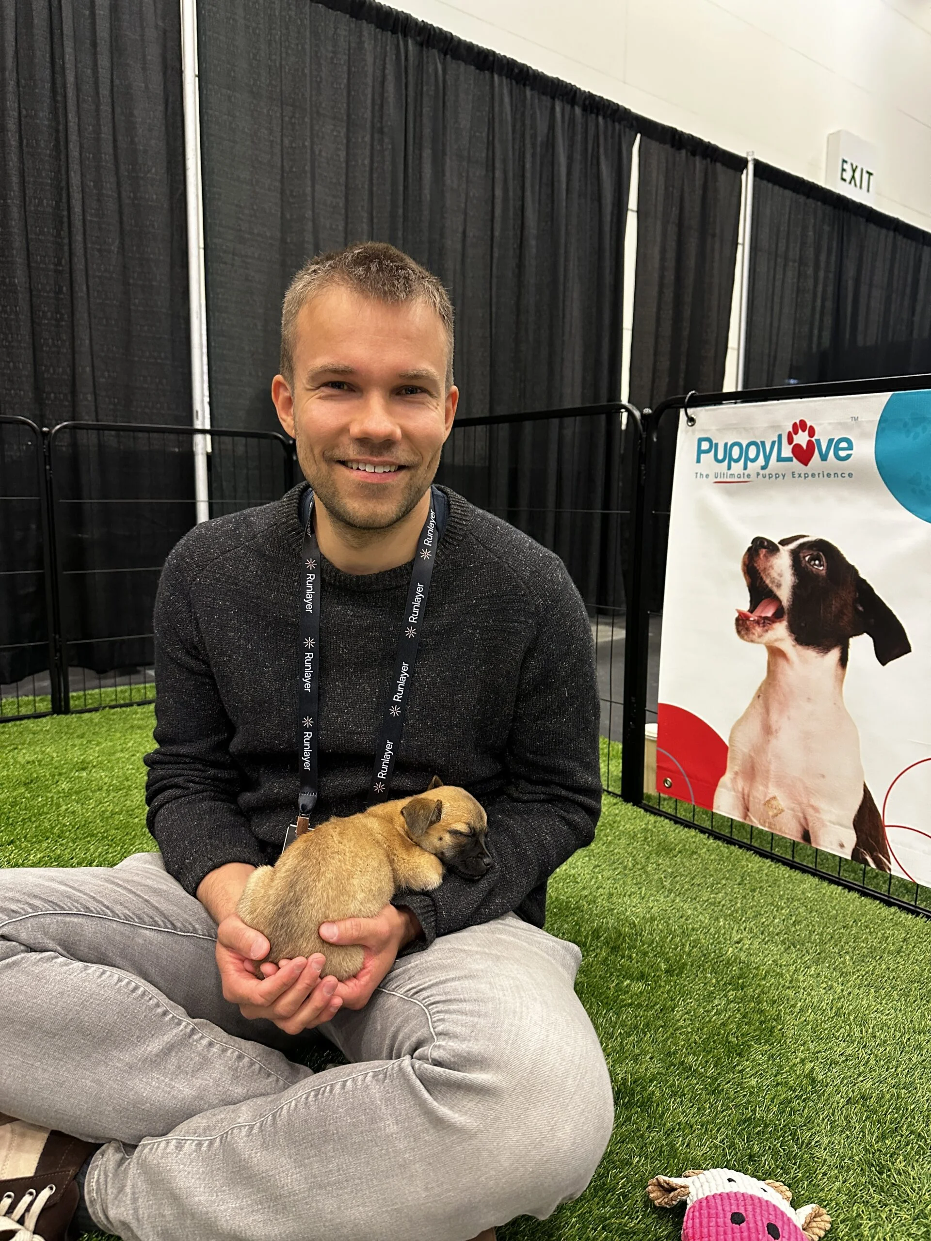 A smiling man with short brown hair wearing a dark grey long-sleeve sweater sitting cross-legged on green artificial turf, holding a small tan puppy in both hands.