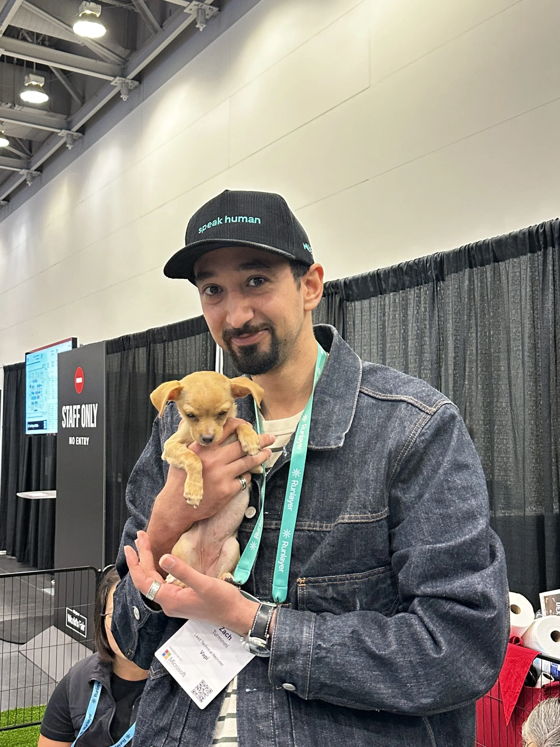 A smiling man wearing a baseball cap with "speak human" text, a denim jacket, and conference lanyards, standing indoors while holding a small tan puppy in both hands.