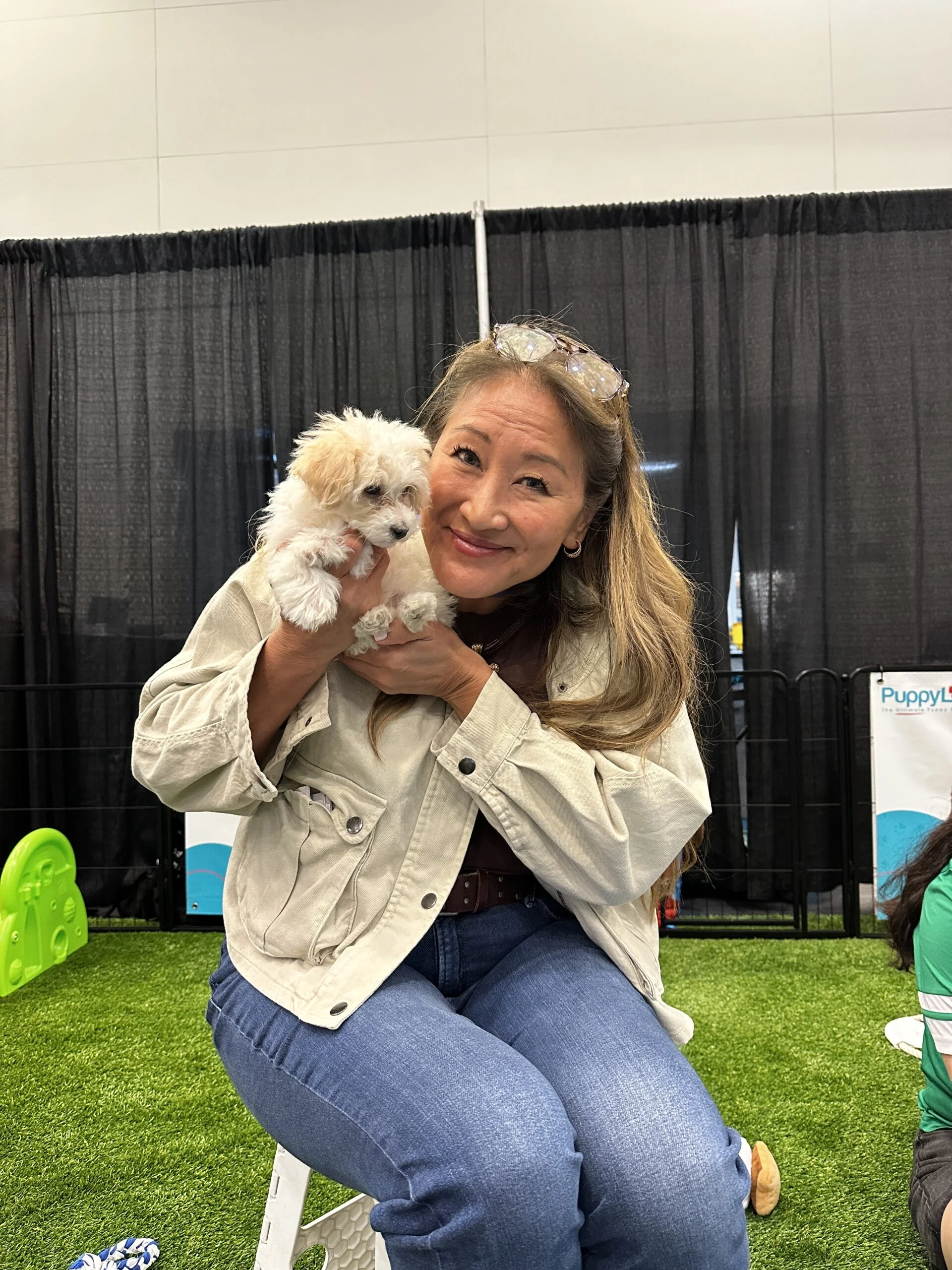 A smiling woman with long highlighted hair wearing a light beige jacket and blue jeans, sitting on artificial turf while holding a small, fluffy white puppy in her arms.
