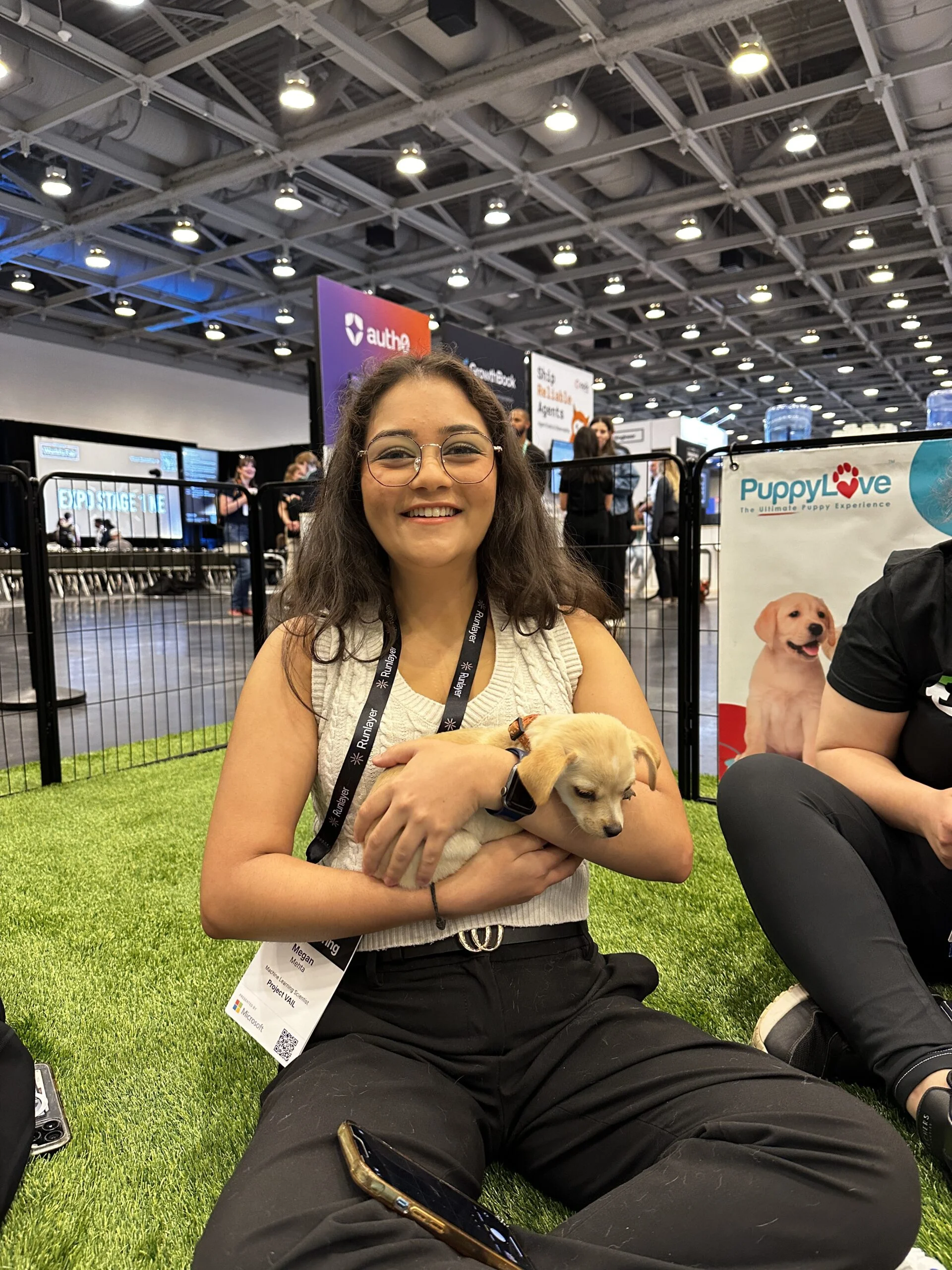 A smiling woman with glasses, dark curly hair, and a beige top sitting cross-legged on green artificial turf while cradling a small tan puppy in her arms.