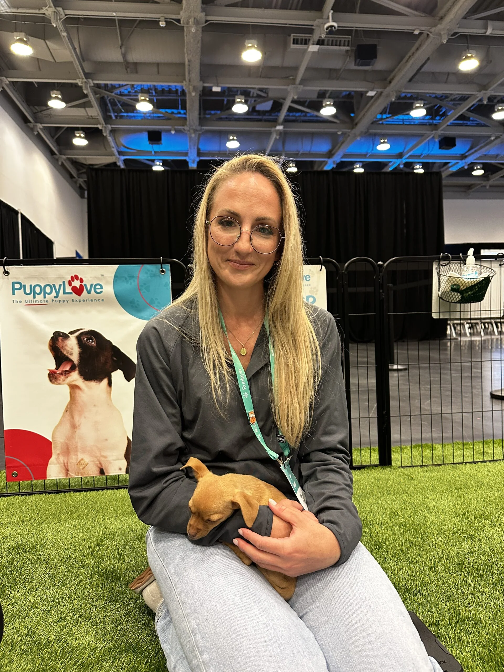 A blonde woman with glasses wearing a grey sweater and white pants kneeling on green artificial turf while cradling a small brown puppy in her hands.