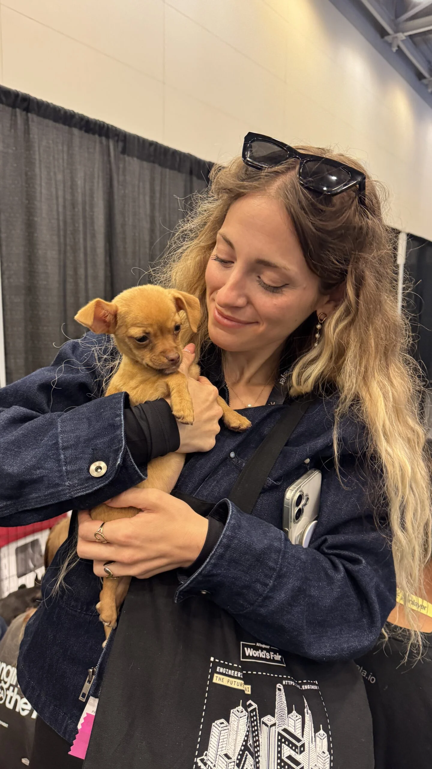 A smiling woman with long wavy blonde hair and sunglasses resting on her head, wearing a dark jacket, holding a small brown puppy in her arms indoors.