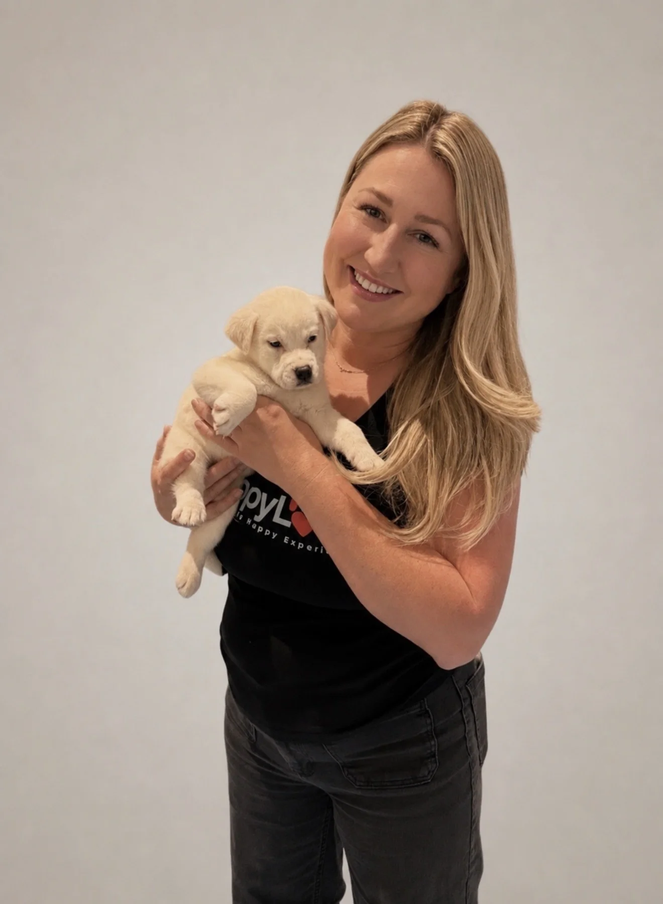 Blonde woman named jessica haro wearing a black puppy love shirt, smiling while holding a small cream-colored puppy in las vegas