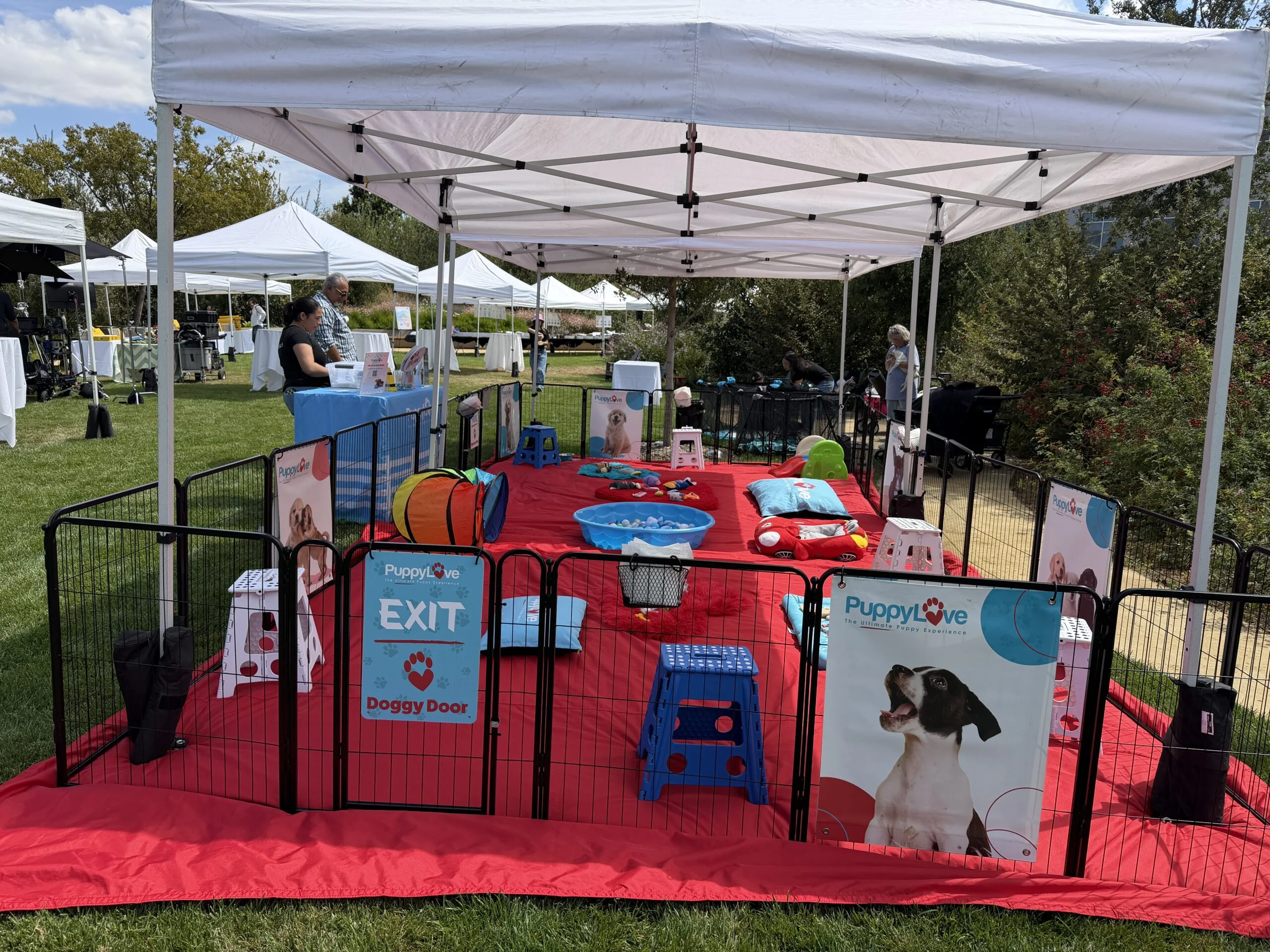 An outdoor puppy play pen with a red tarp, puppy love signs, an "exit doggy door" gate, toys, and white event tents on a grassy lawn.