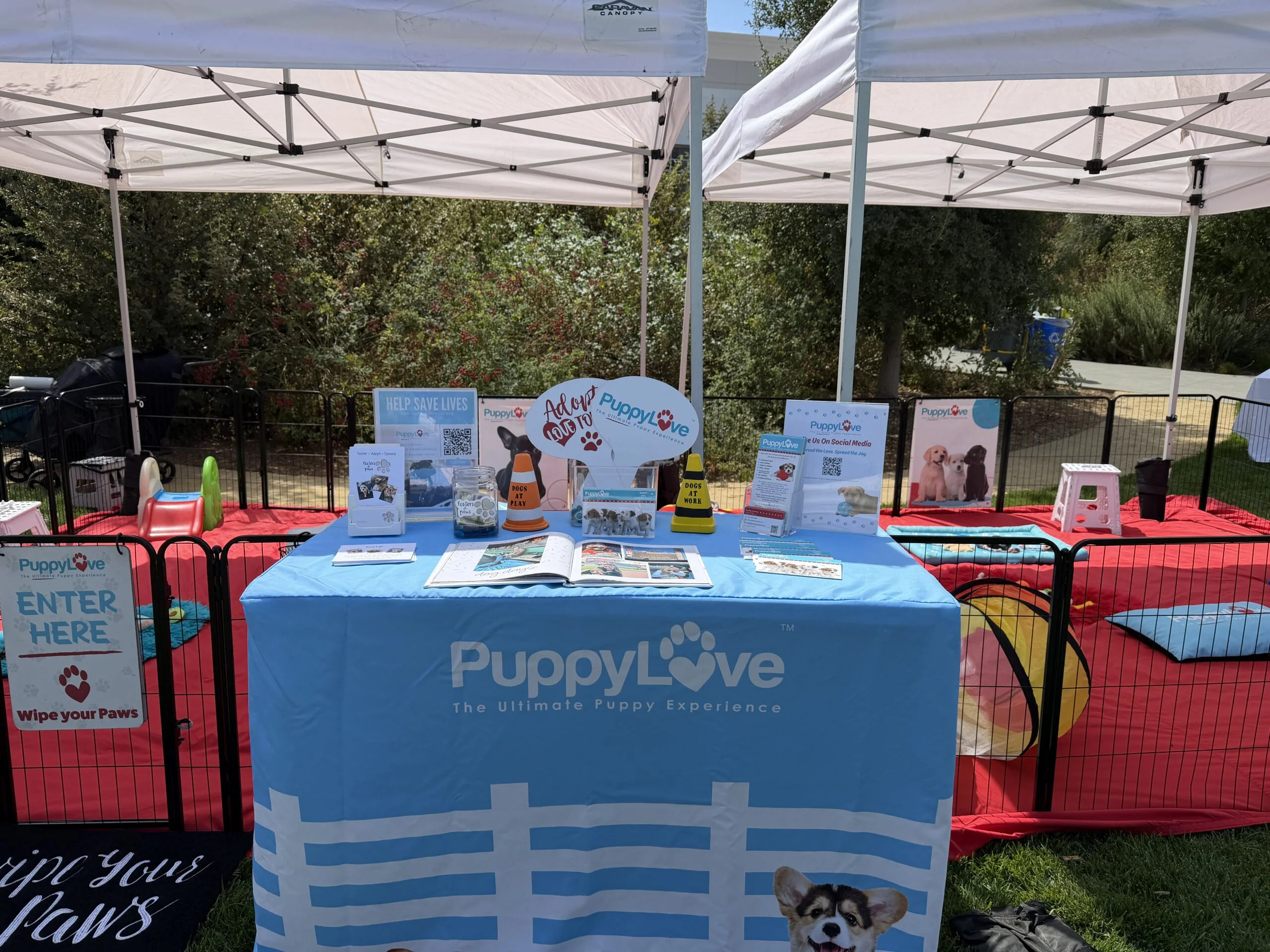 An outdoor puppy love event booth set up with branded blue tablecloths, informational displays, and surrounding playpens on a grassy lawn.