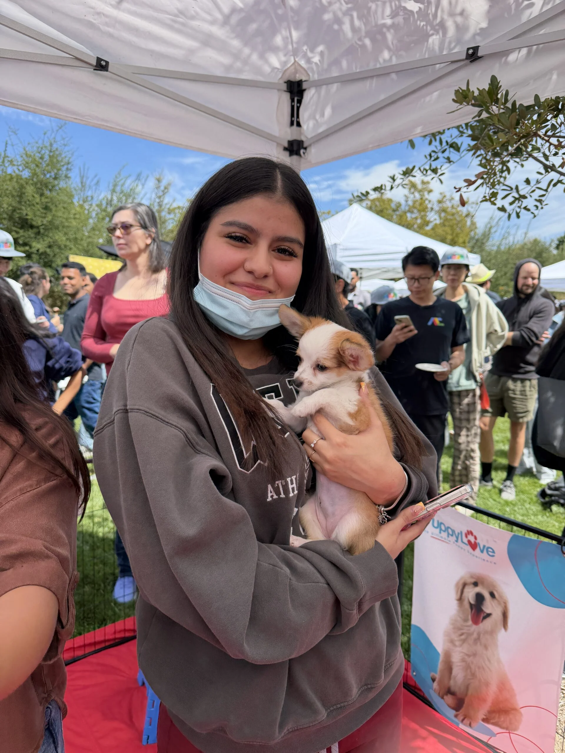 A woman wearing a face mask and grey hoodie smiling under an outdoor tent while holding a tiny white rescue puppy at a google san jose event.