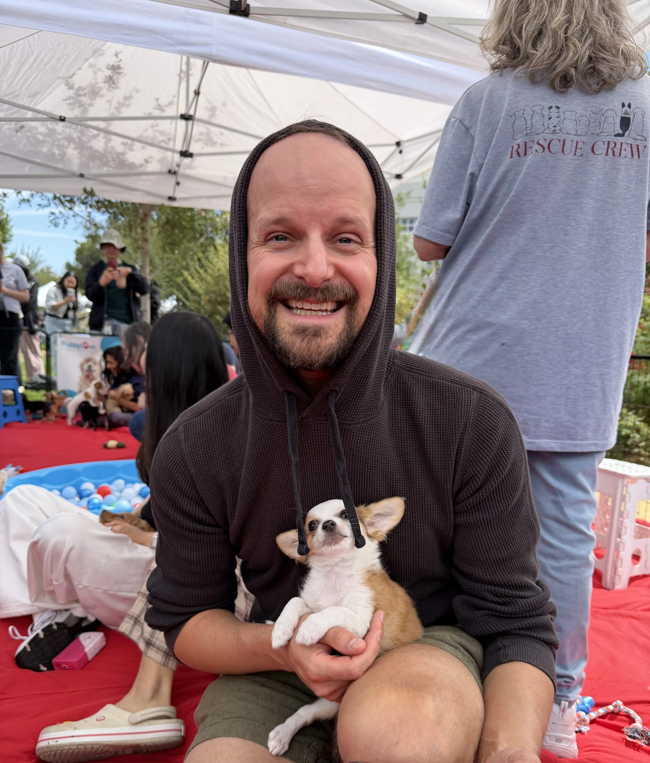 A man in a black hoodie sitting outdoors on a red tarp, smiling while holding a tiny white chihuahua puppy in his lap at a google san jose event.
