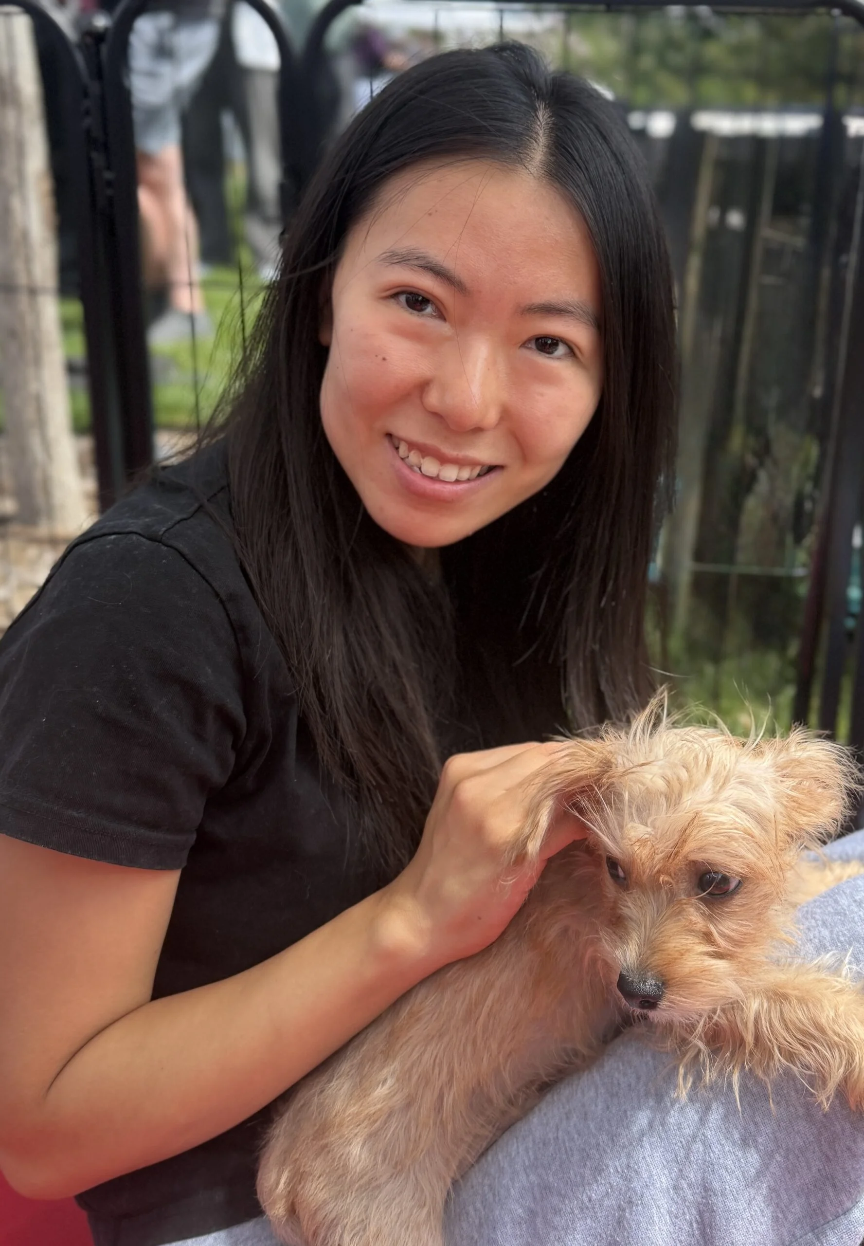 A smiling woman with dark hair sitting outdoors, gently petting a small scruffy tan dog in her lap.