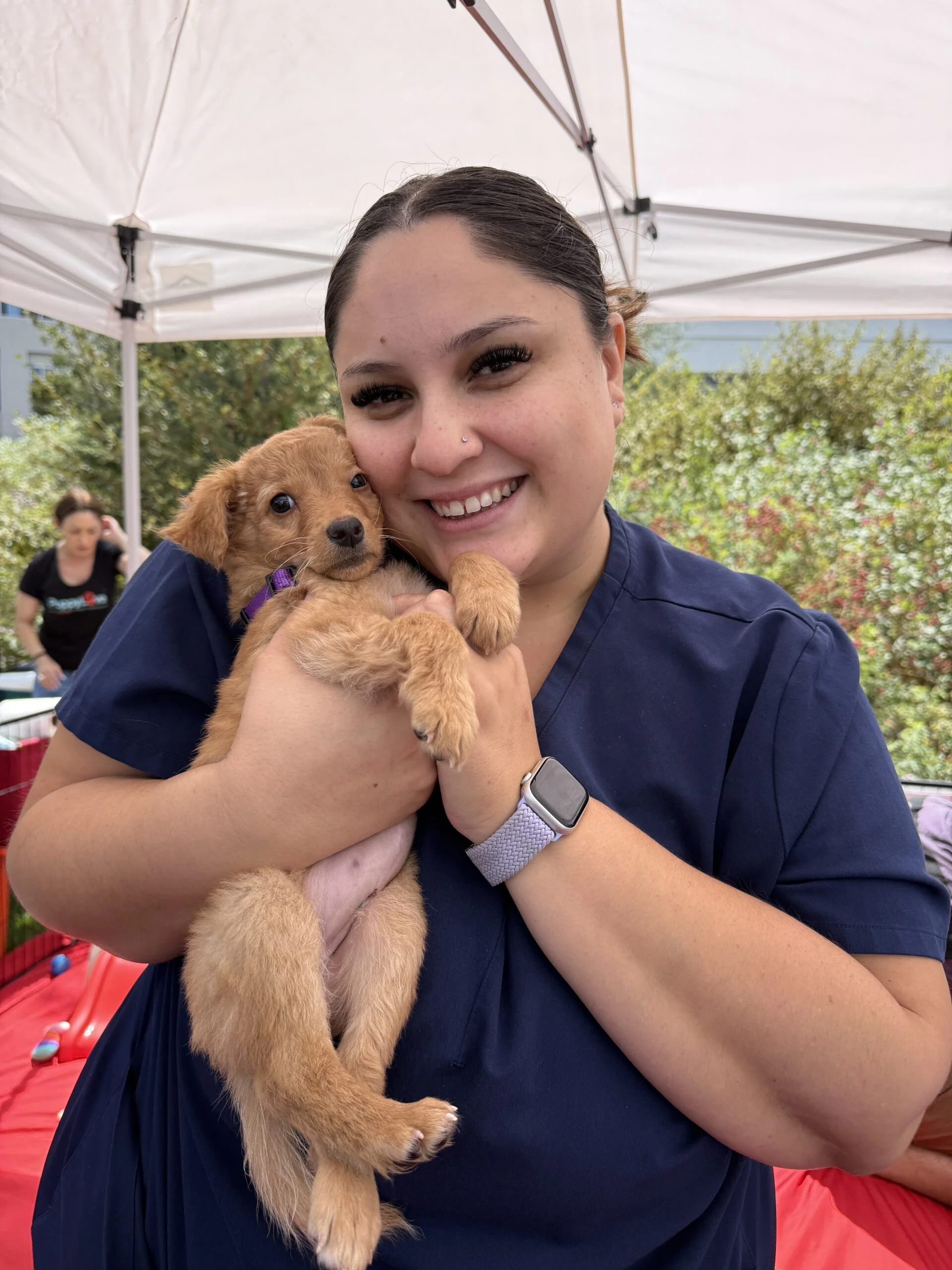 A woman in dark scrub-style clothing smiling outdoors while holding a fluffy, light brown rescue puppy.