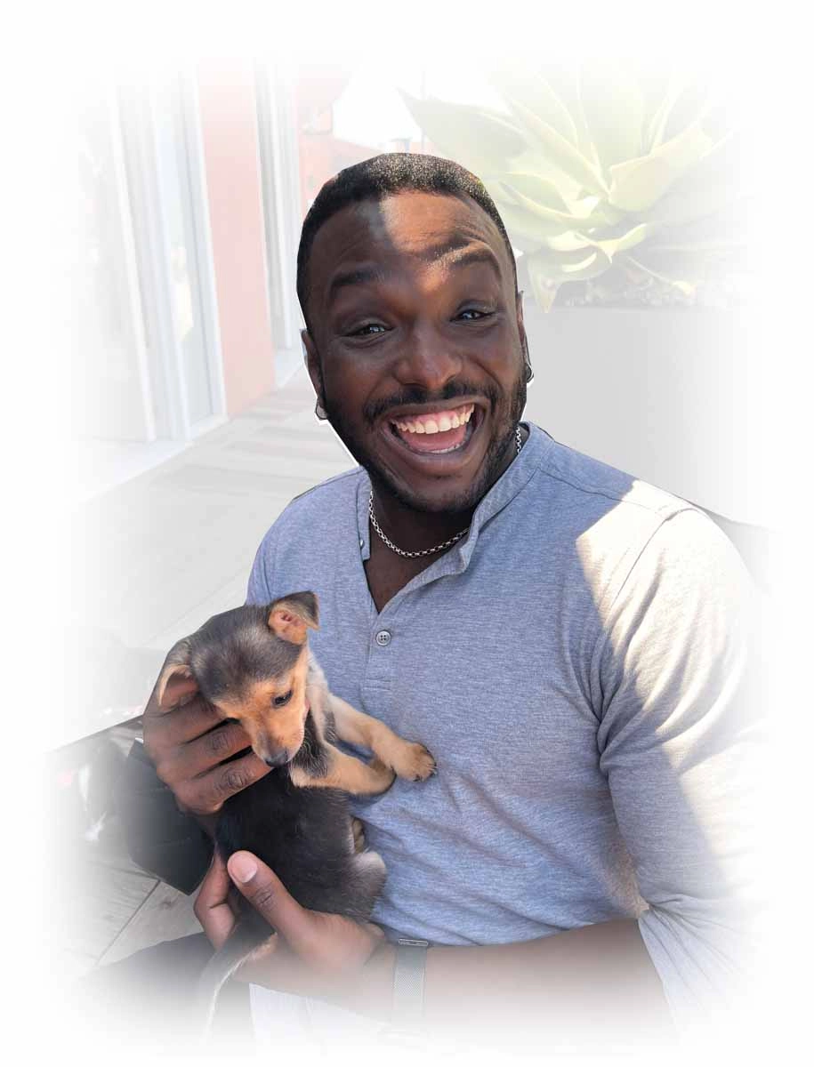 A smiling man holding a small brown puppy outdoors in front of a building.